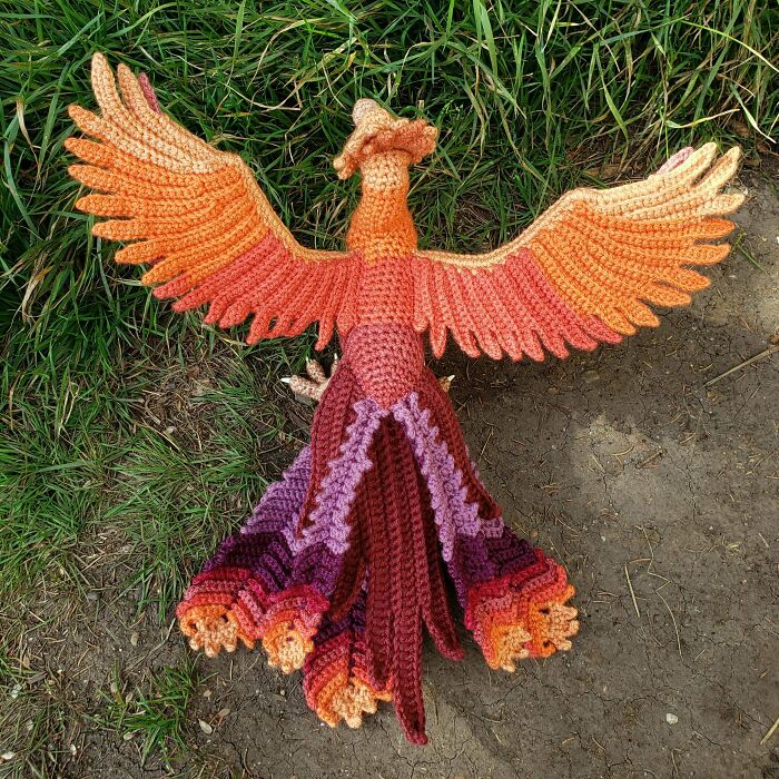 Crochet masterpiece of a colorful phoenix with detailed wings and tail displayed outdoors on grass and dirt.