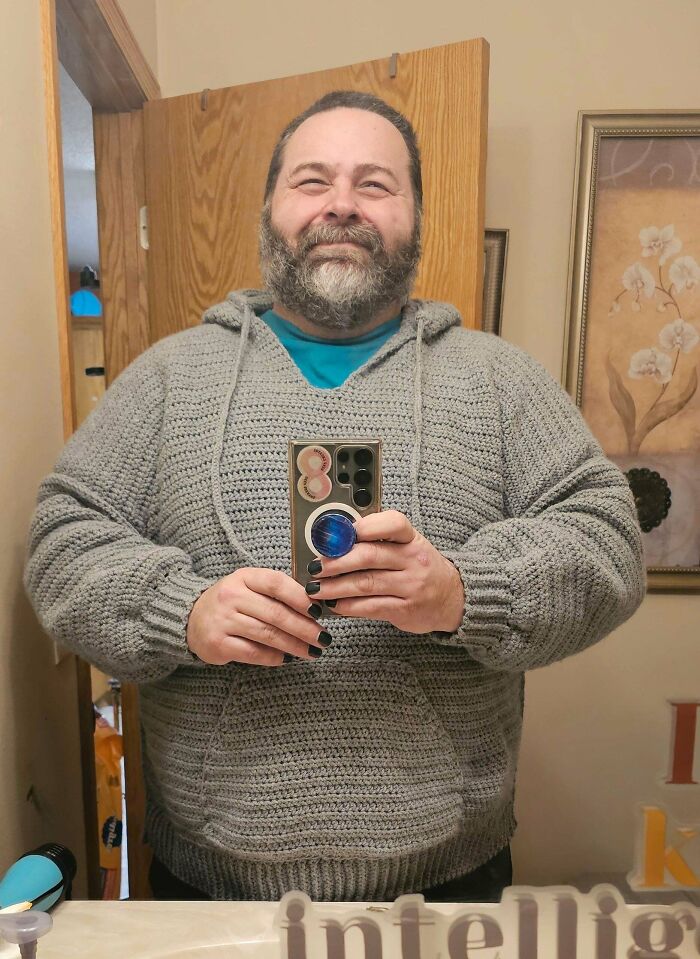 Man smiling and taking a mirror selfie wearing a handmade gray crochet hoodie showcasing crochet masterpieces.