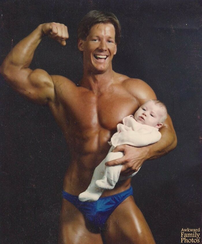 Muscular man flexing while holding a baby in a vintage awkward family photo with a dark background.