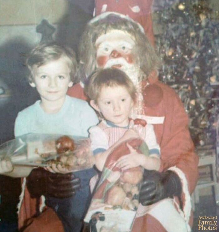 Two children sitting with a person dressed as Santa Claus in an awkward family photo that also feels sweet by the Christmas tree.