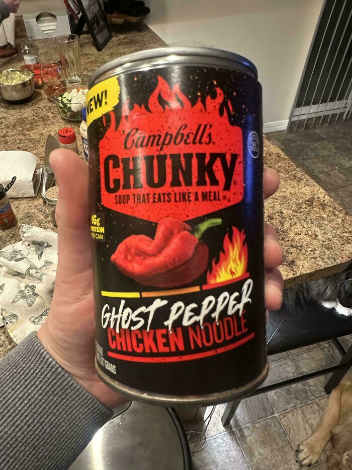 Hand holding a can of Campbell's Chunky Ghost Pepper Chicken Noodle soup on a kitchen countertop with scattered items.