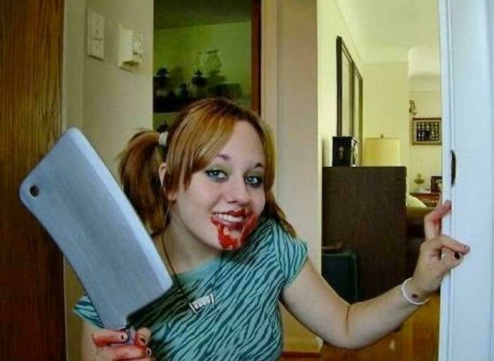 Young woman with blood makeup on face holding a large cleaver, posing in a room showcasing a regrettable past moment.