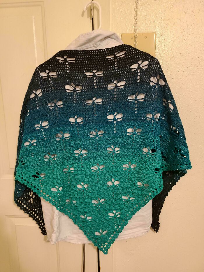 Gradient turquoise and black crochet shawl with dragonfly patterns hanging on a white hanger and door.