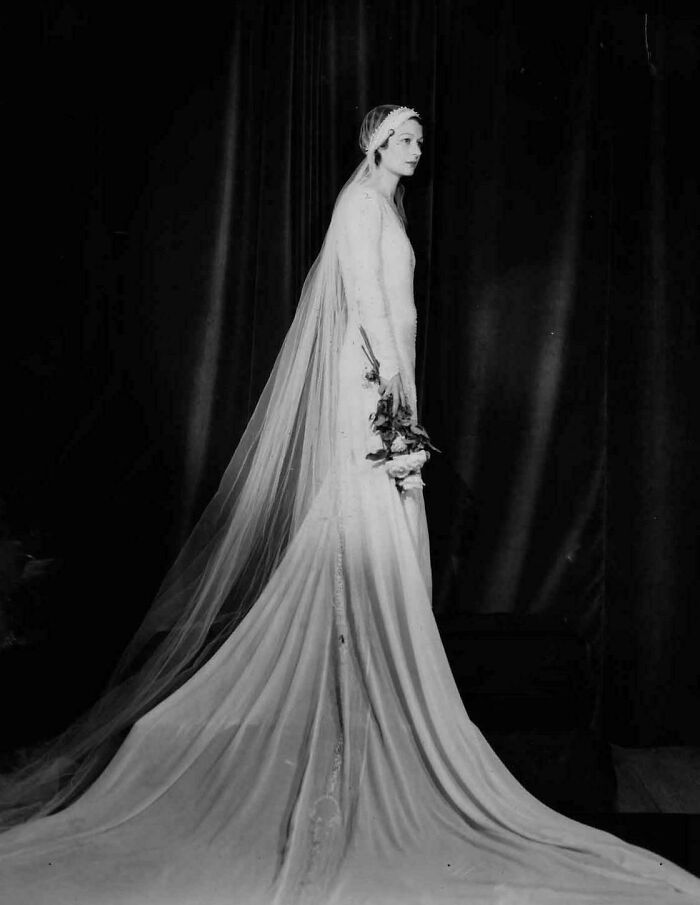 Black and white old image of a bride wearing a long flowing gown and veil holding flowers in a vintage setting.
