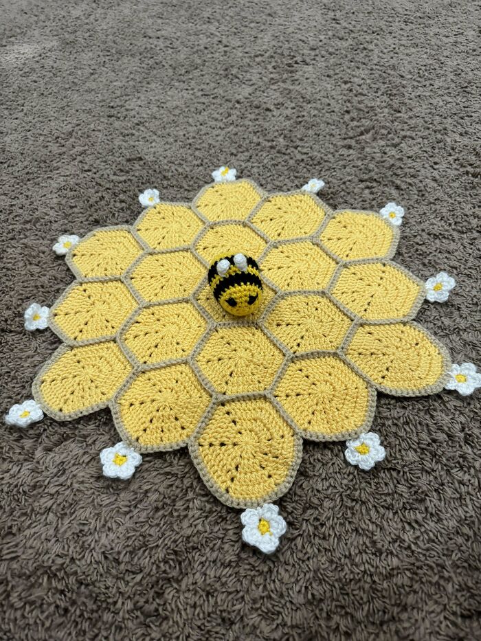 Yellow crochet hexagons arranged like a honeycomb with white flowers and a small crocheted bee in the center on a beige carpet.