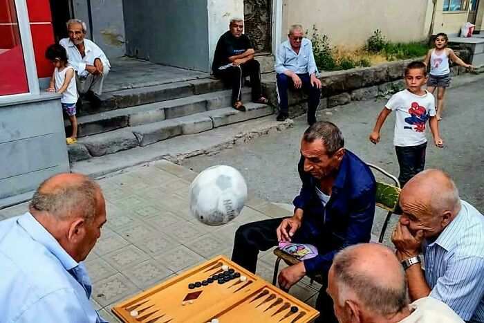 Men playing board game on street, capturing an accidental Renaissance scene with a soccer ball mid-air.