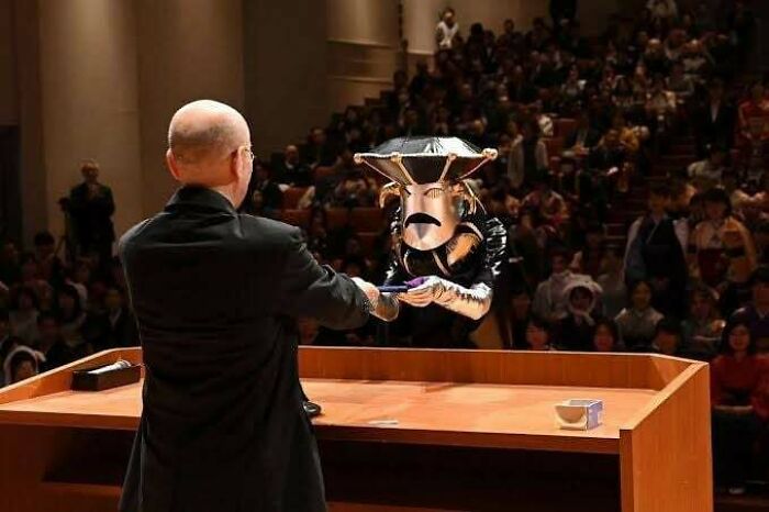 A man in a suit, facing a person in a unique costume on stage, in front of a large audience; fascinating facts scenario.