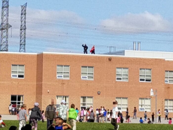 Children playing outside a school while two people stand on the rooftop, evoking old-school memories.