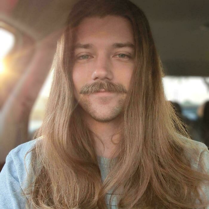 Man with fierceflow beautiful hair and a styled mustache sitting in a car with sunlight streaming in from the side.