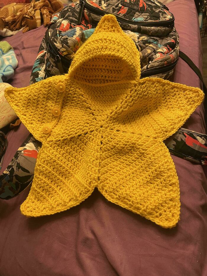 Yellow crochet star-shaped baby blanket with buttons, displayed on a bed with a patterned backpack in the background.