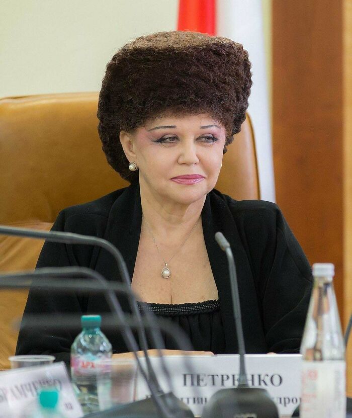 A woman with a large, unfortunate haircut resembling a fur hat, wearing a black top and a pearl necklace.
