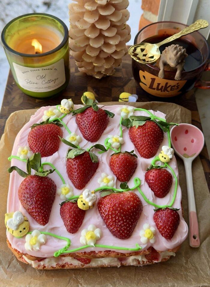 Square strawberry cake with pink icing, decorated with edible bees and flowers, a candle, and a mug on a wooden board.