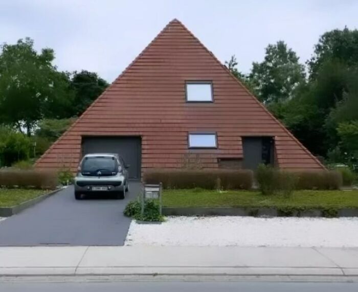 Pyramid-shaped house with brick facade, two small windows, and a car in the driveway, showcasing hilariously bad architecture.