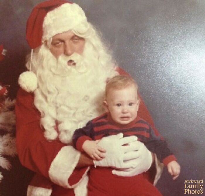 Santa Claus holding a crying baby in a striped outfit, capturing an awkward family photo moment that feels sweet.