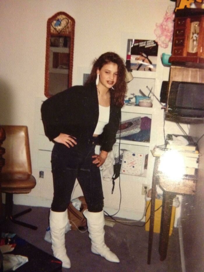 Woman dressed in 80s fashion with black jacket, leggings, and white boots, standing in a cluttered room.