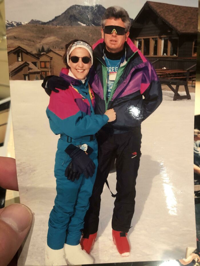 Two people in colorful 80s fashion ski outfits, wearing sunglasses, posing on snow with mountains and a cabin in the background.