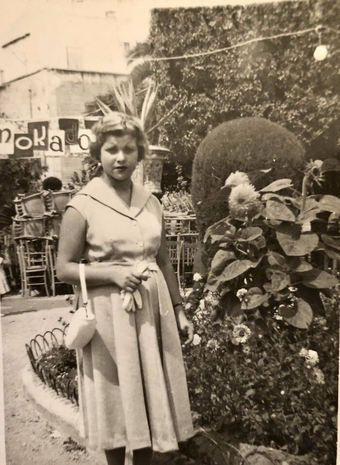 Vintage photo of a woman in a dress holding gloves, standing in a garden with chairs and a hedge in the background.