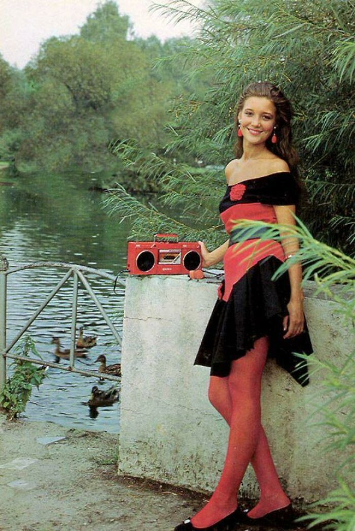 Woman in 80s fashion with red tights and black dress, standing by a river, holding a red boombox.