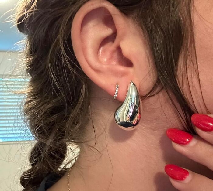 Silver teardrop earring and diamond stud on woman's ear, red nails visible.