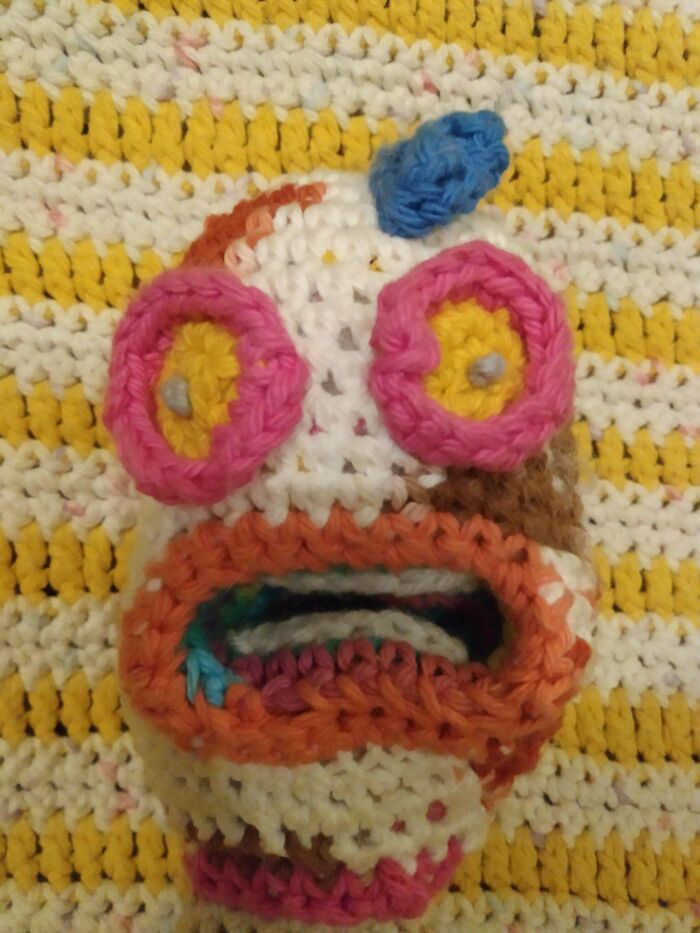 Close-up of a colorful crochet piece with large eyes and an open mouth on a yellow and white crochet background.