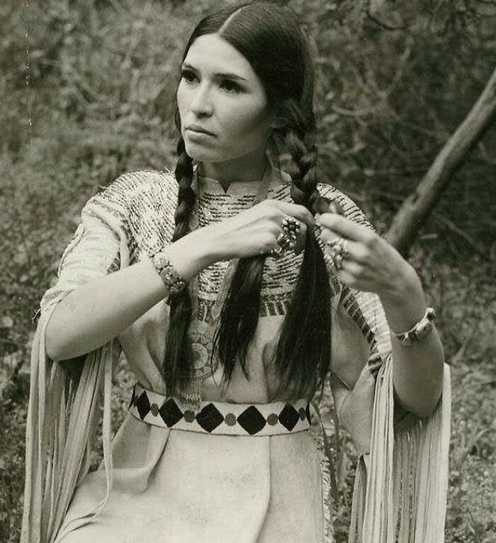 A woman braiding her hair, dressed in traditional attire, with intricate jewelry, depicting a moment from the past.