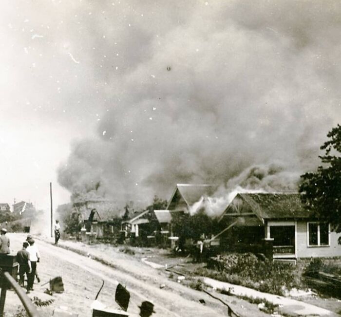 Historic scene of houses engulfed in smoke, with people observing the smoky aftermath on a dirt road.