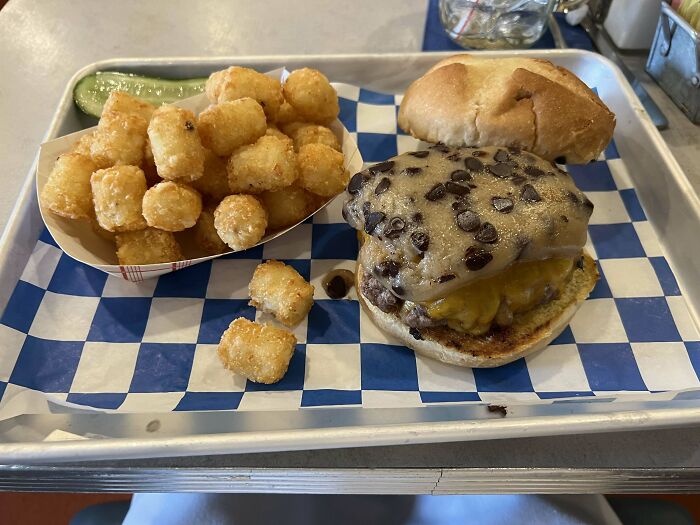 Chocolate chip cookie burger with tater tots and a pickle on a tray, showcasing unusual and silly food dishes.