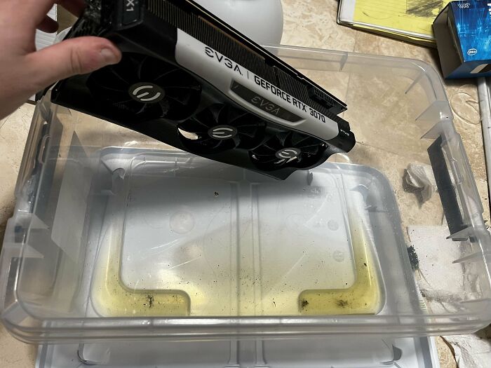 Hand holding EVGA GeForce RTX 3070 graphics card above a plastic container with liquid, illustrating tech support work challenges.