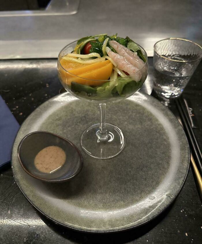 Salad served in a cocktail glass on a large plate with dressing and glass of water showcasing ridiculous restaurant food presentation.