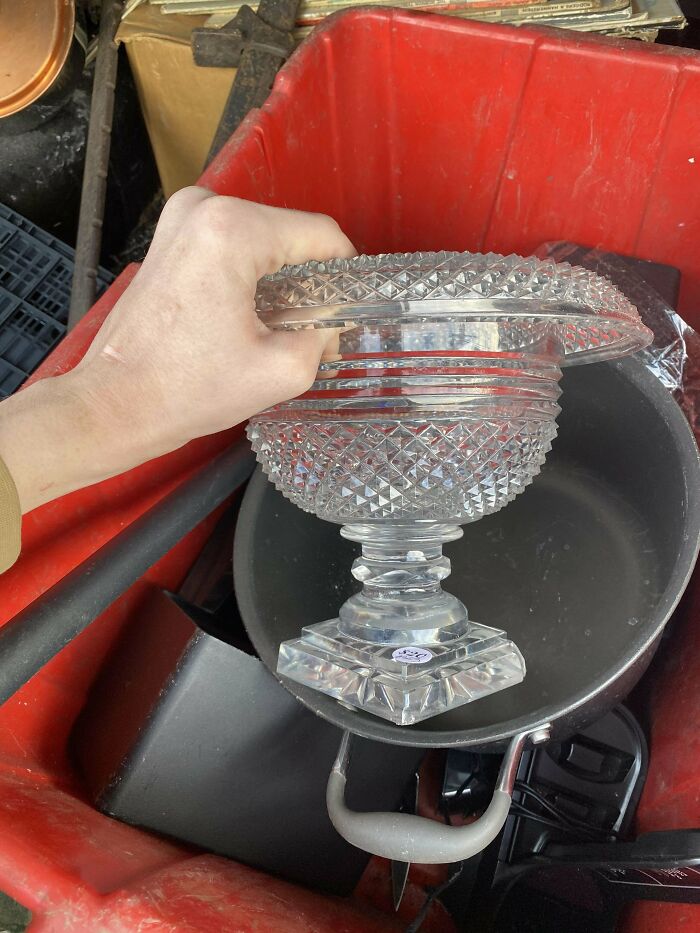 Hand holding a crystal bowl found while dumpster diving in a red container, next to a frying pan.