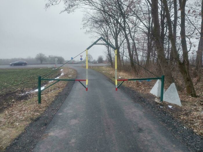 Mildly interesting pic of a path blocked by a colorful geometric metal structure among trees, with a road in the background.