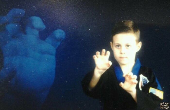Young boy in martial arts uniform posing awkwardly for a family photo with a large ghostly hand in the background, sweetly imperfect.