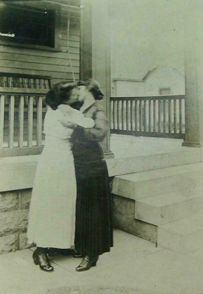 Victorian era women sharing a kiss on a porch, both wearing long skirts and boots, capturing intimacy in a historical context.