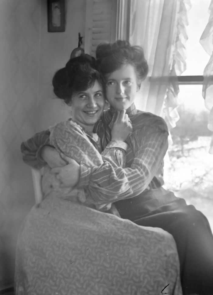 Two women in Victorian-era attire, smiling and embracing by a sunlit window.