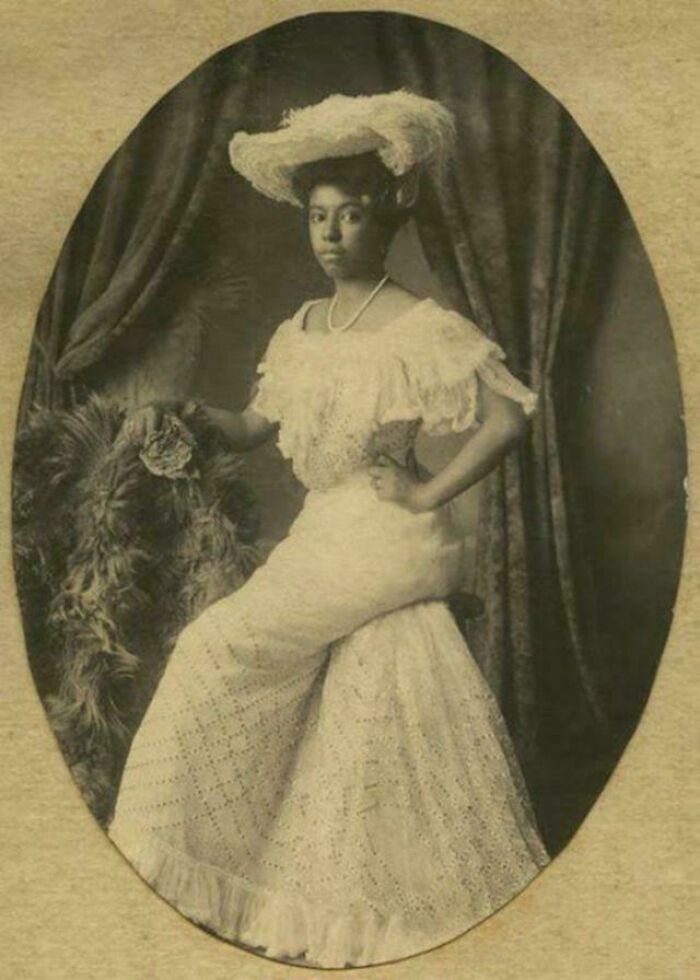 Victorian Era woman in elegant lace dress and hat, posing with hand on hip in a vintage portrait setting.