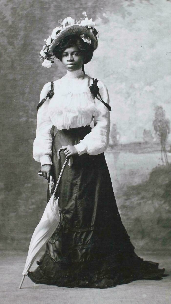 Victorian era woman in a floral hat and long skirt holding a parasol, showcasing period fashion.