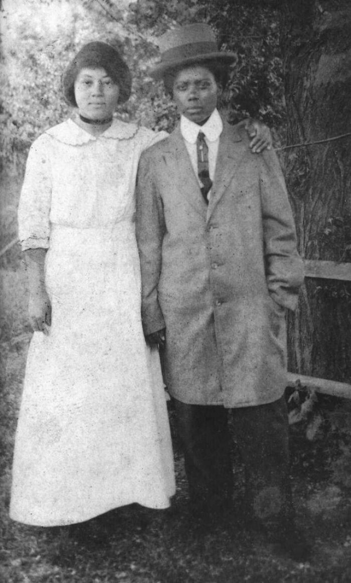 Victorian Era couple posing outdoors, woman in white dress and man in suit with hat, arm around each other.