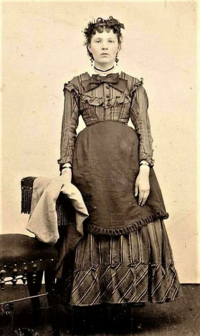 Victorian era woman in striped dress, standing beside a chair.