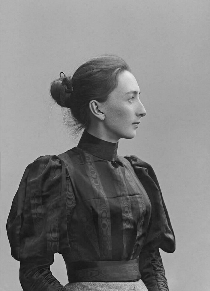 A woman in Victorian-era attire, featuring a high-neck blouse and elaborate sleeves, with hair in a bun.