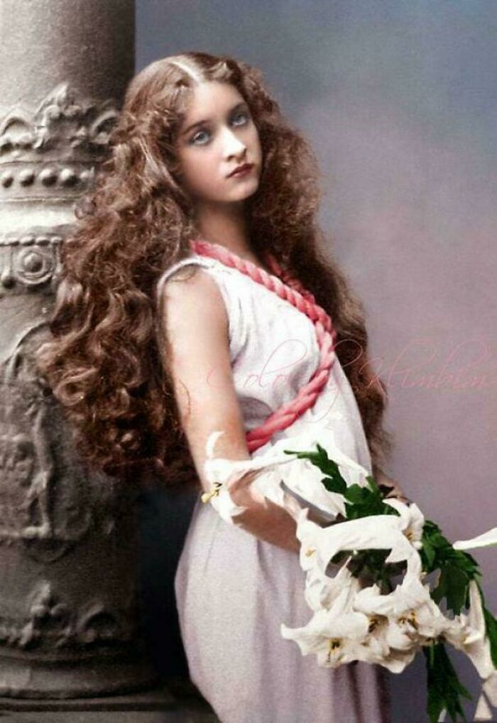 Victorian era woman in elegant attire holding white lilies, with long curly hair, standing by an ornate pillar.