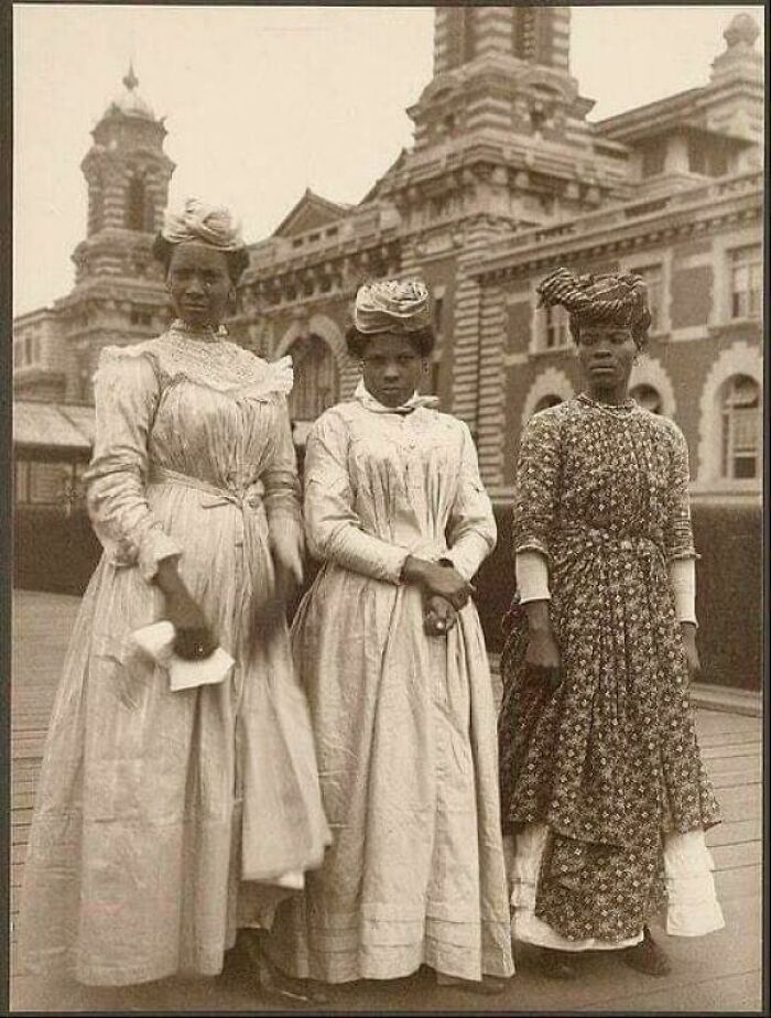 Victorian era women in period dresses posing outdoors in front of a historical building.