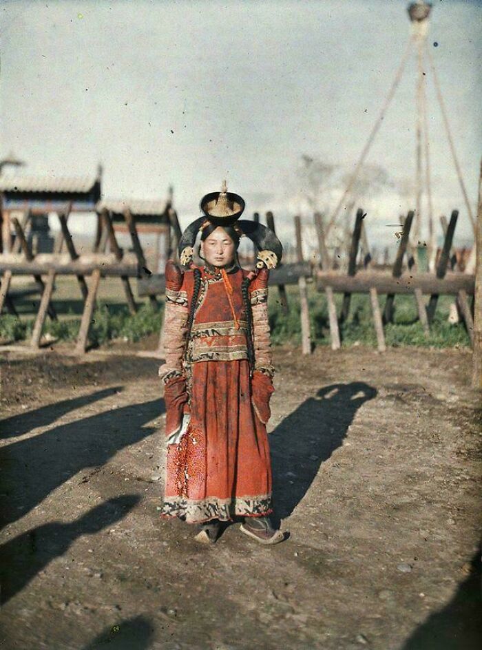A woman in traditional Victorian-era attire stands on a dirt path, with wooden structures in the background.