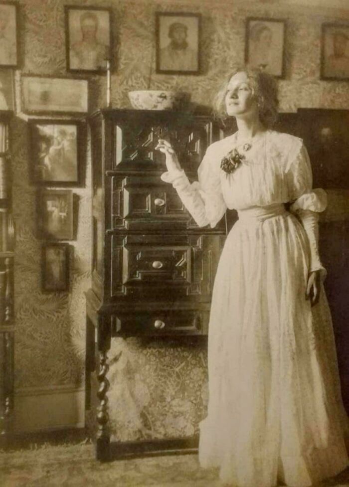 Victorian era woman in elegant dress, standing in a vintage room with framed portraits on the walls.