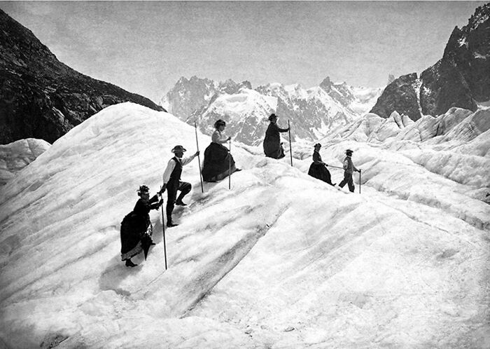 Victorian era explorers in formal attire climbing a snowy glacier with mountains in the background.