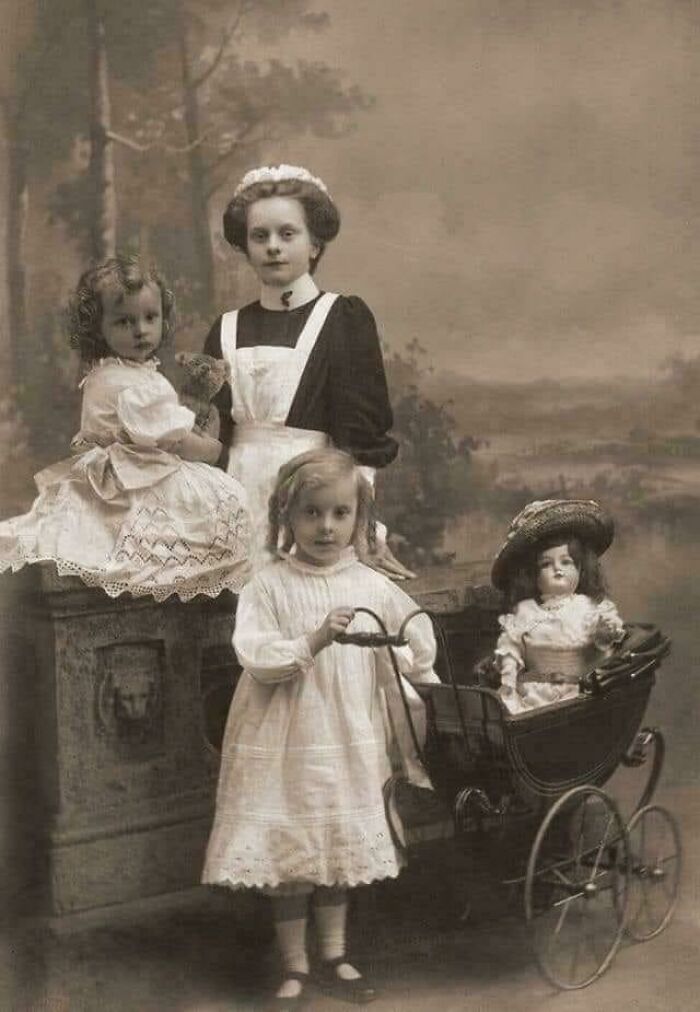 Victorian era children with a nanny in period clothing, one child holding a doll in a pram, posed in a studio setting.