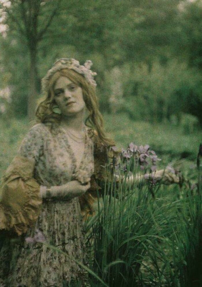 Victorian era woman in a floral dress standing in a lush garden, holding purple flowers.
