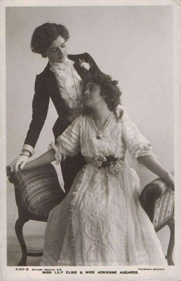 Victorian era women in period fashion, one standing, the other seated on an ornate chair, in a classic pose.