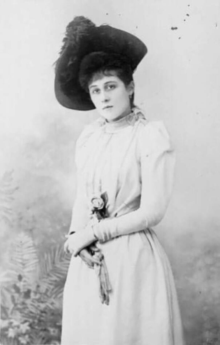 Victorian era woman in a long dress and large hat, standing with a gentle expression.