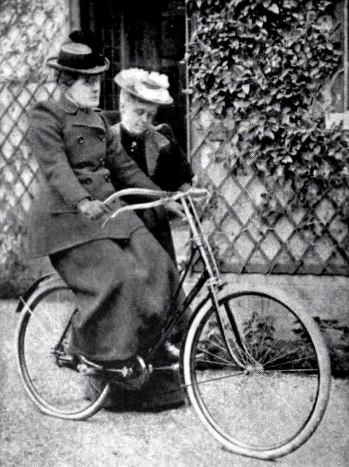 Victorian era women in hats and coats riding a vintage bicycle beside a vine-covered wall.