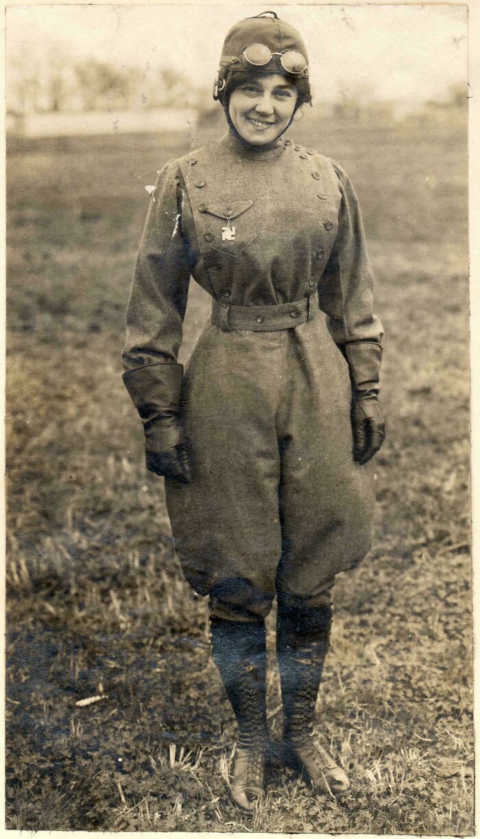 Victorian era woman in aviation gear, wearing a flight cap and goggles, standing outdoors with a smile.